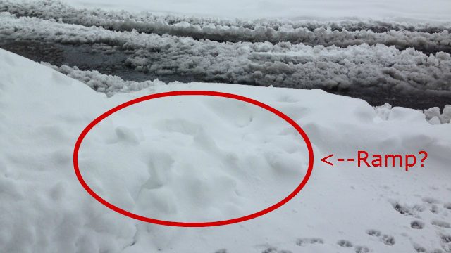 snow covered curb with a red circle and text pointing to an area where there may be a ramp but it is impossible to tell because of the snow
