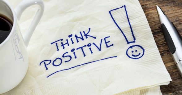 Napkin with "think positive!" written on it. On top of the napkin is a mug of coffee and next ot the napkin is a pen.