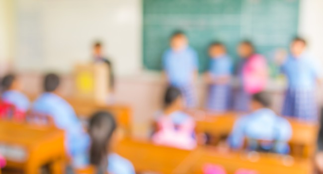 A blurred photo of students in a classroom, sitting in desks and standing in front of a chalkboard.