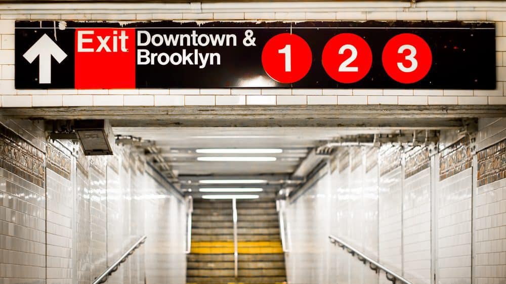 Photo of hallway leading to steep stairs. Above is a sign that says "Exit, Downtown & Brooklyn - 1, 2, 3."
