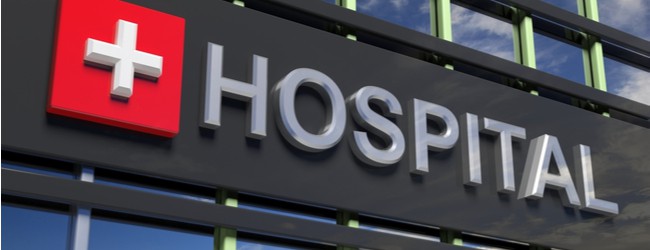 A close-up of a black sign on a building that says "Hospital" in gray letters. Next to the text is a red square with a gray cross symbol on it.