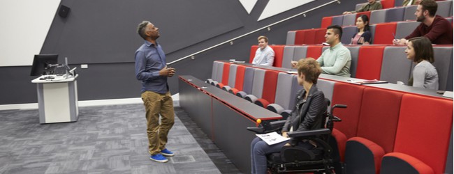 A group of students in a lecture hall, sitting with several open seats in between each student. A student in a power chair sits at the bottom of the stadium-style lecture hall seating, separate from peers.