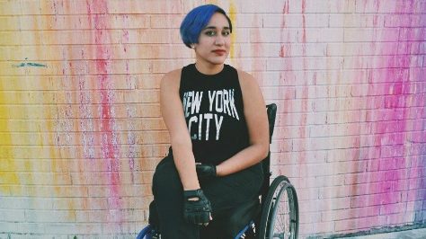 Photo of Annie Segarra with blue hair in a black outfit. Sitting in a wheelchair in front of a pastel rainbow splattered wall.