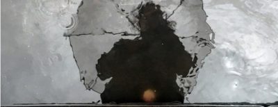 A darkened, unclear reflection of the top half of a person holding an umbrella, looking down at a grayish puddle of water.