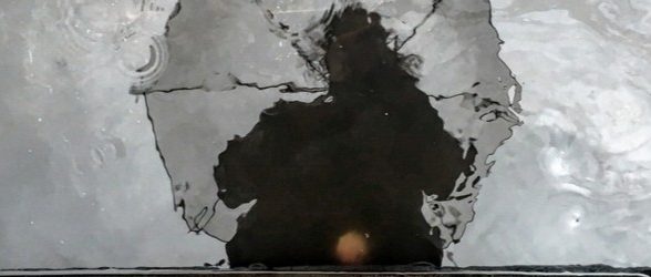 A darkened, unclear reflection of the top half of a person holding an umbrella, looking down at a grayish puddle of water.
