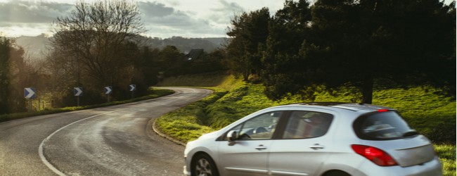 A silver car driving on an open road surrounded by trees, greenery, and a grayish sky.