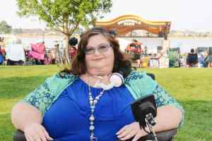Photo of Carrie Ann Lucas Carrie wearing blue and teal cat eye glasses, curly hair, a blue dress, long necklace, and a multicolored blue and teal jacket. Carrie's trach and ventilator tubing is pictured, along with portions of her wheelchair controller. 