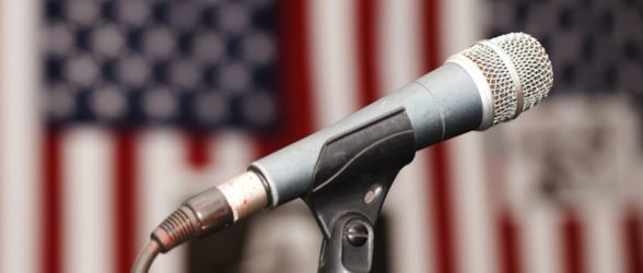 A photo of a microphone on a stand in front of blurred American flags in the background.