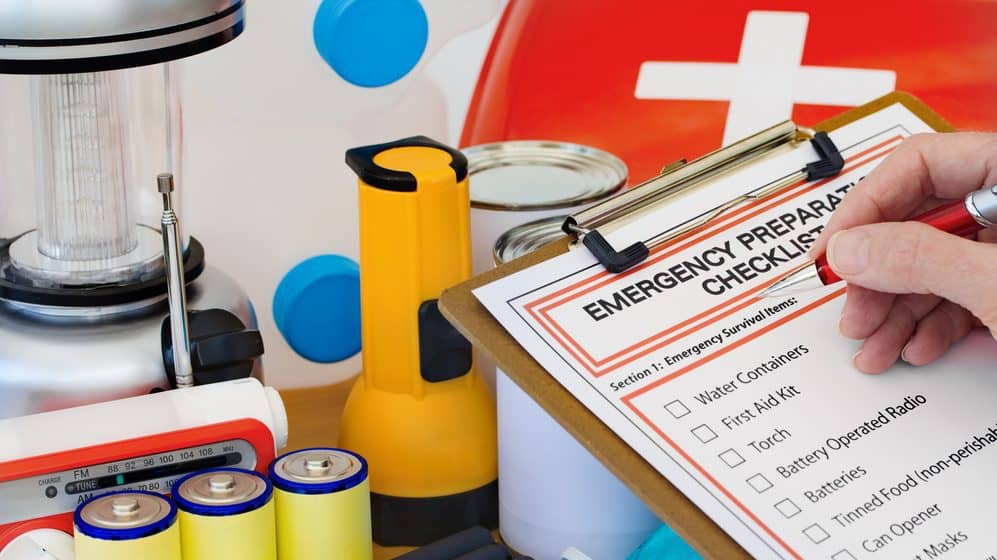 Emergency supplies including flashlight, radio, and waterbottle. A person is writing on an emergency preparedness checklist attached to a clipboard