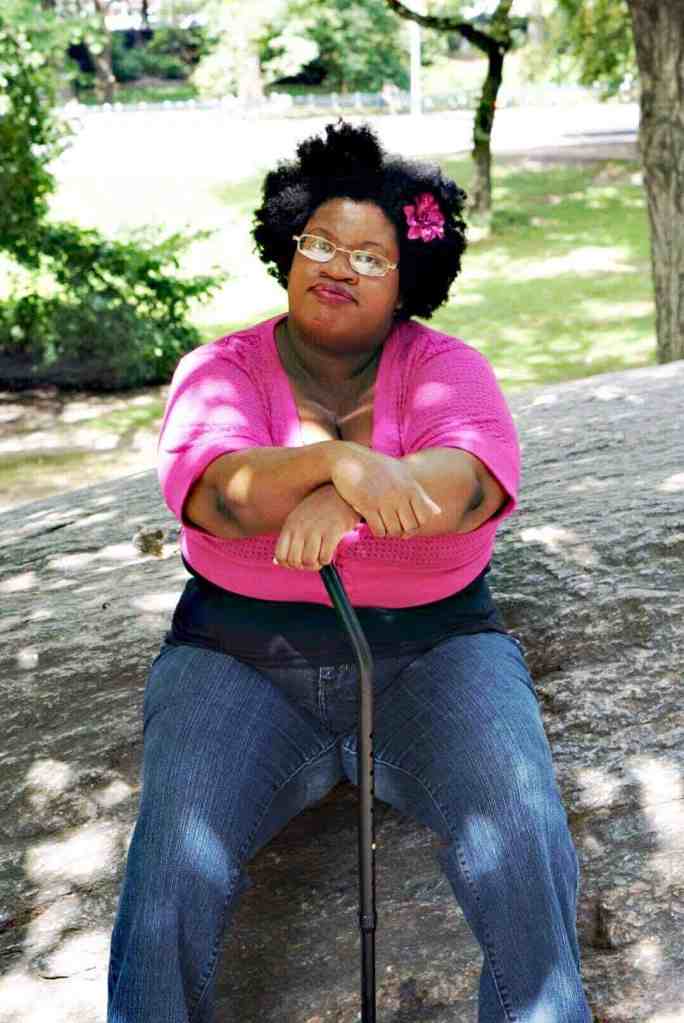 Outdoors, day time. Denarii, a proudly fat, brown-skinned Black woman, is wearing a pink flower in her afro, a black tank top with a pink half-sweater over it, and dark denim jeans. She's sitting on a large gray rock, resting her hands on her black cane. She's facing the camera, not smiling. In the background are the green trees and grass of Central Park.