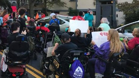 Large group of ADAPT members, many using various mobility devices, holding signs during an action.
