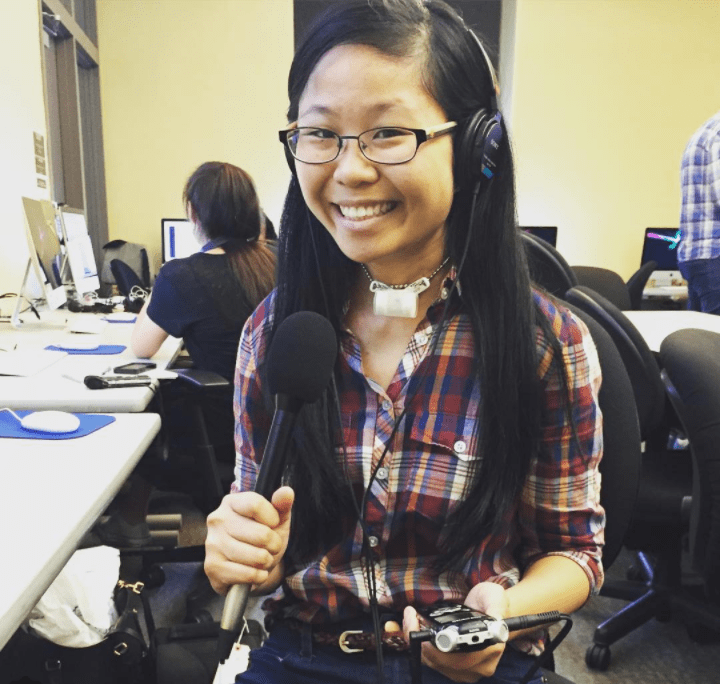 A Chinese American woman with glasses and a tracheostomy tube smiles at the camera, wearing a red and blue plaid shirt and blue jeans with a thin belt. She is wearing a black audio headset and holding up a padded microphone in her right hand and a black recorder in her left hand. She's sitting in a classroom with Mac computers and students sitting or bustling in the background.