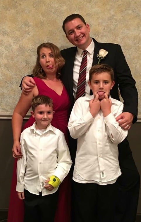 A photo of a family, a mom, dad, and two young boys, all dressed up. The mom is wearing a red dress, the father is wearing a suit and tie, and the two boys are in white button-down shirts and black pants.