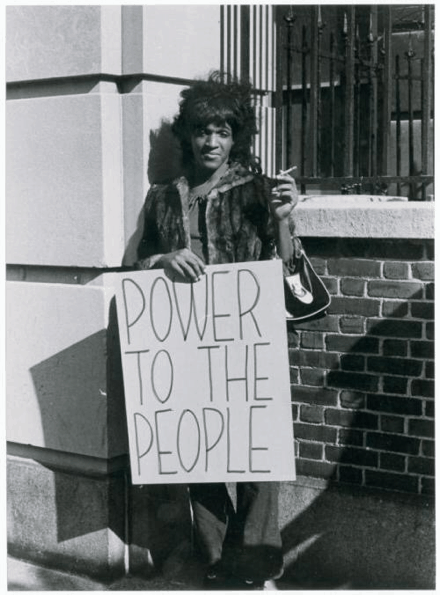 Marsha P. Johnson, a Black trans woman, stands with a sign reading “POWER TO THE PEOPLE.” She is wearing a fur coat and holding a cigarette.