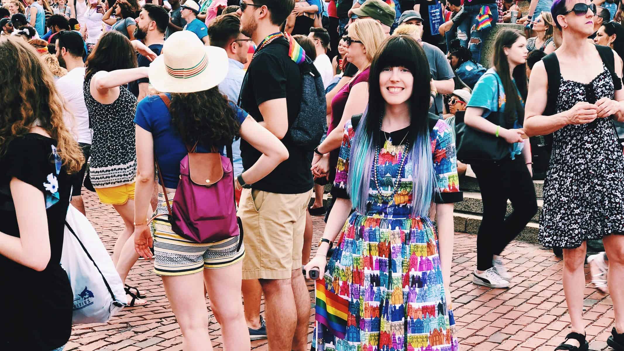 Alaina, a white thin femme person, standing in front of a crowd at Pride in Boston. She is wearing a dress with rainbow castles all over it. I have long hair that is dark brown on top and a mix of blues and purples on the bottom. She is standing with hert lavender cane with a rainbow flag attached to it, and smiling.