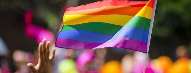 A supporting hand waves in front of a rainbow flag flying on the sidelines of a summer gay pride parade