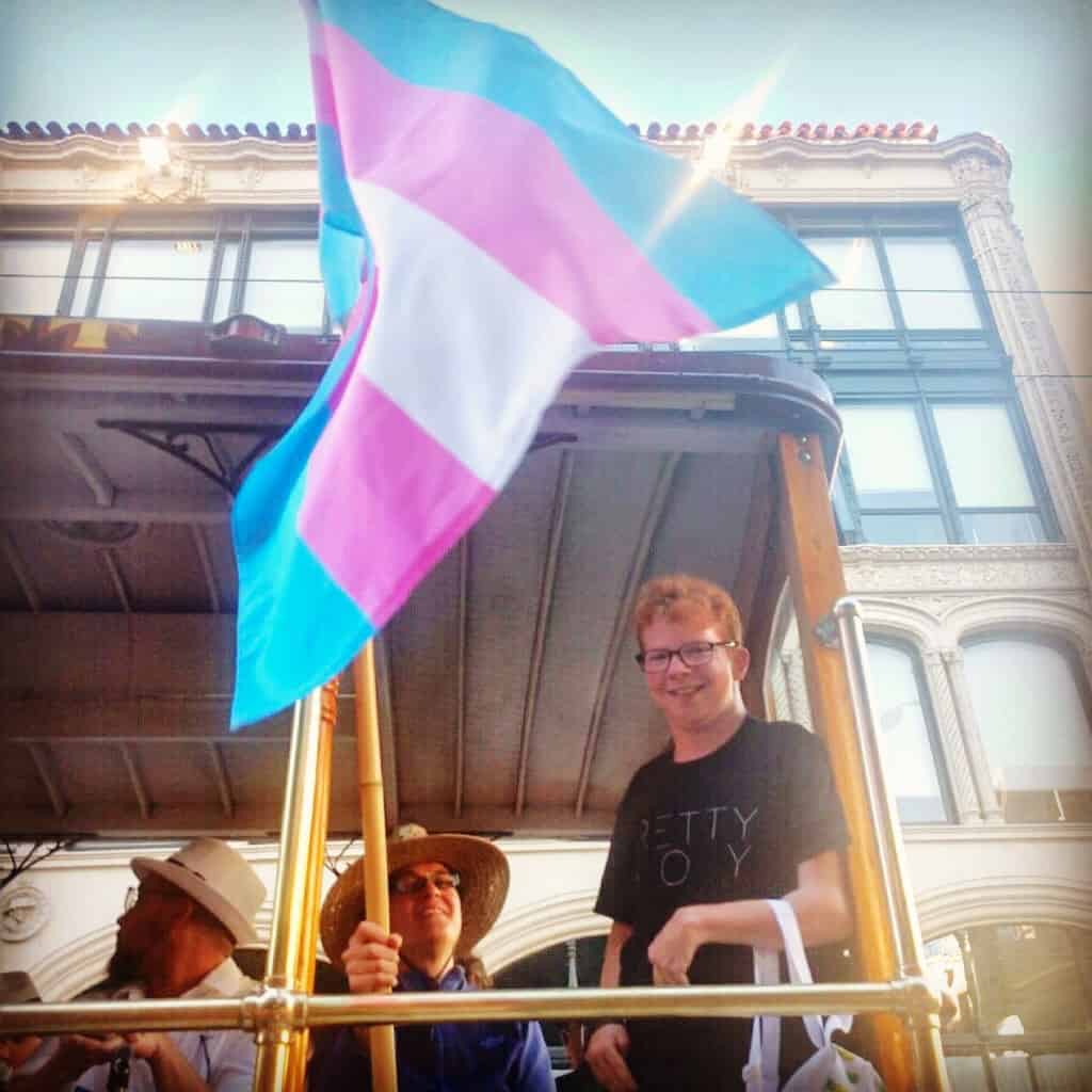 Christian, a white trans man, is on a trolley. He is smiling. Next to him, someone is holding the Trans Pride flag.