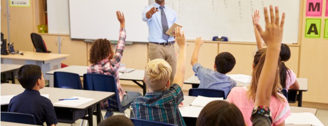 A teacher at the front of the class pointing who a student whose hand is raised. There is a group of students sitting in front of the teacher at their desks.