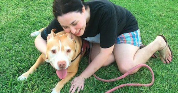 Photo of Brijana, wearing shorts and a tshirt, sitting on the grass, hugging her pit bull, whose tongue is sticking out.