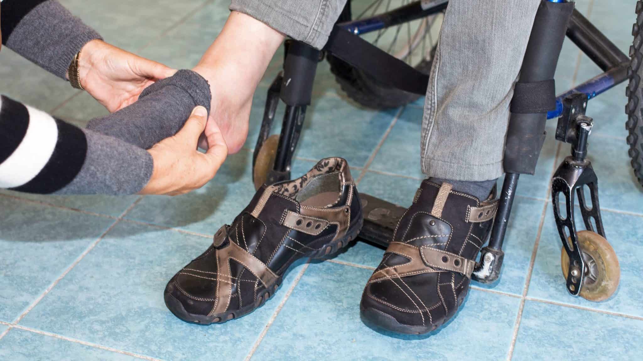 Photo showing a person kneeling down, assisting a person sitting in a wheelchair with putting on a sock.