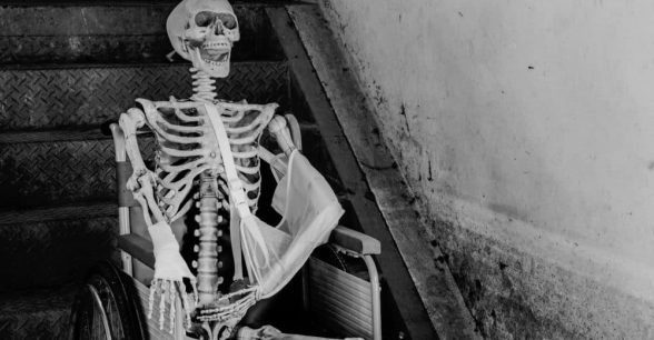 Black and white photo of skeleton sitting in a hospital-style wheelchair in front of stairs.