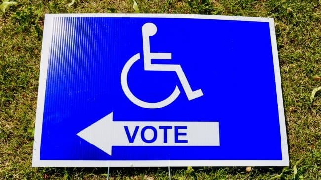 A bright blue sign mounted in grass that has an accessible icon and an arrow that says vote on it.