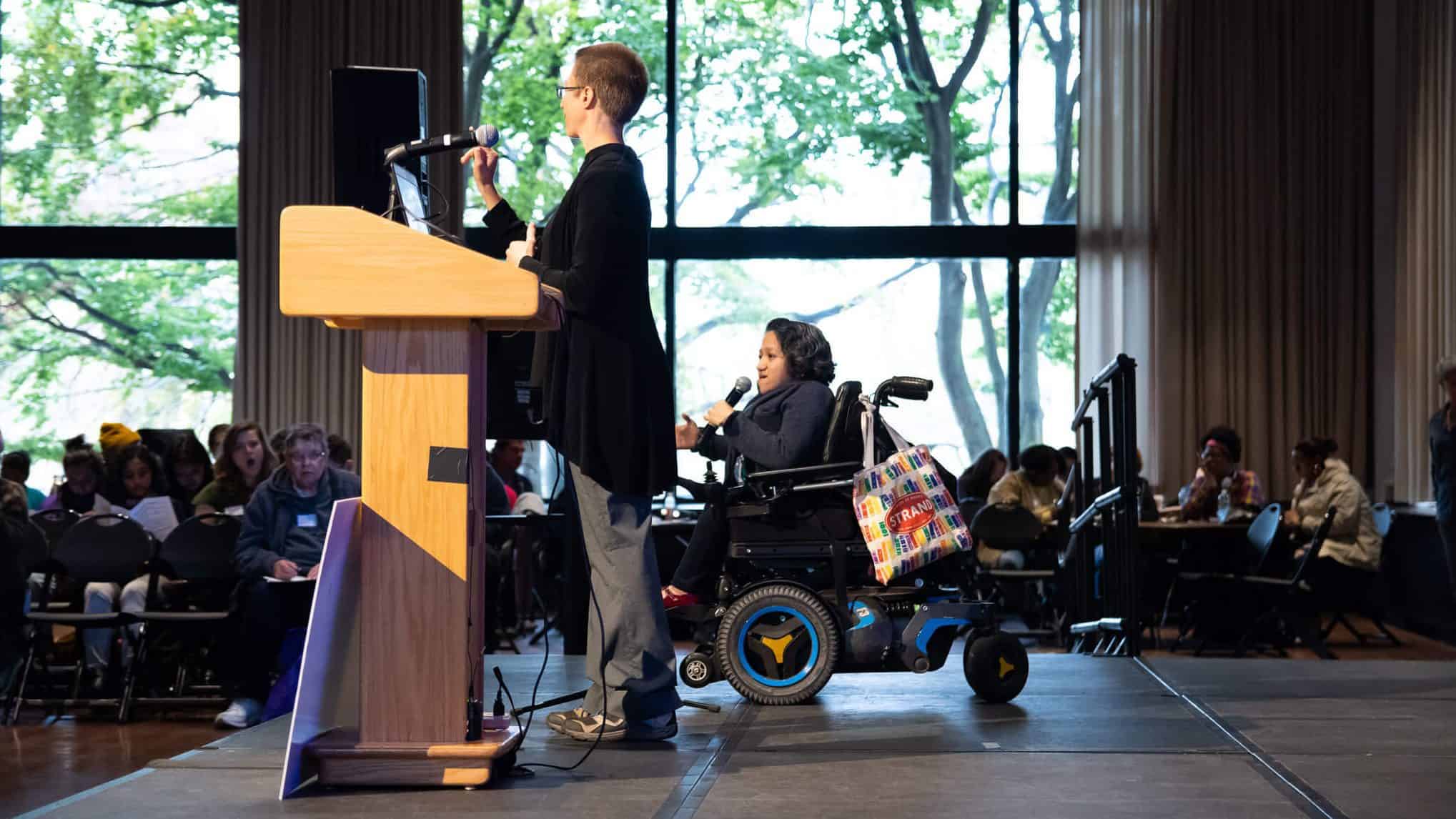 Sandy Ho sits in her power chair and speaks into a microphone, next to another speaker on a stage in front of an audience.