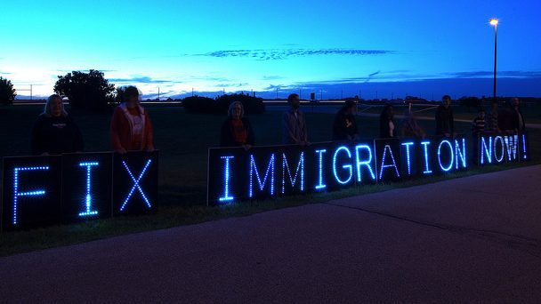 Lit up letters spelling "Fix Immigration now" against a dark blue sky with people standing behind the letters.