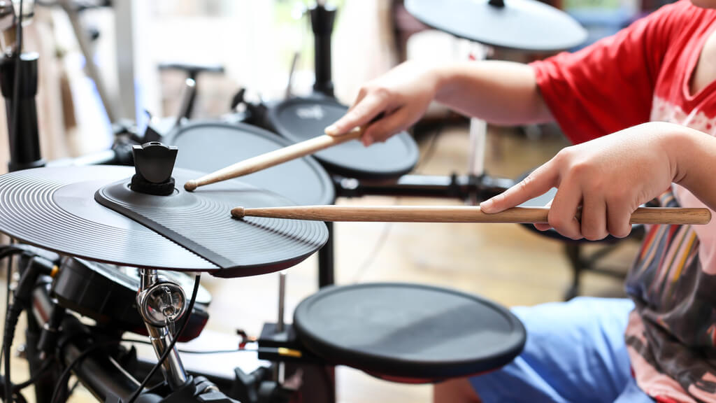 Photo of a young person playing drums. The person's face is not visible, but they are positioned behind the drum set.