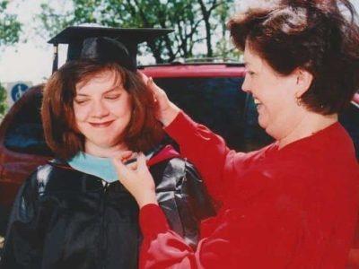 Lisa Ferris in a graduation gown. Her mother is helping her adjust her cap. They are standing outside in front of a red vehicle.