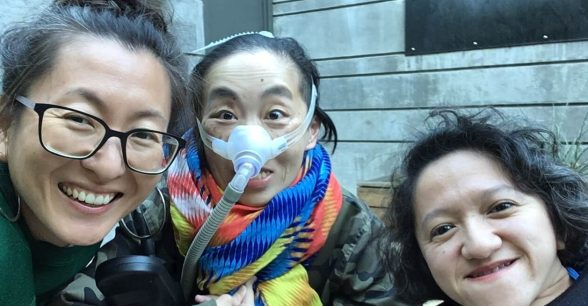 Photo of 3 disabled Asian American women, Mia Mingus, Alice Wong and Sandy Ho (from left to right). Mia is wearing glasses and large hoop earrings. Alice is wearing a brightly colored scarf and an army-camouflage-print jacket. She is wearing a mask over her nose with a tube for her Bi-Pap machine. Sandy has wavy short hair and is wearing a black sweater. Behind them is a concrete wall with a door.