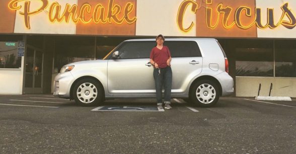 Photo of Christian standing in front of his car, which is parked sideways across accessible spots, in front of a restaurant called Pancake Circus
