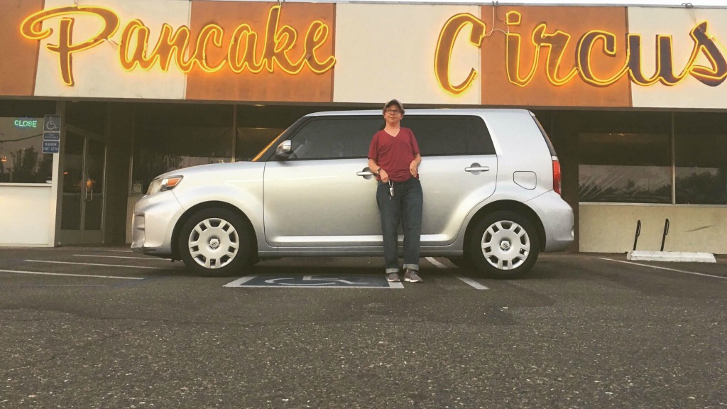 Photo of Christian standing in front of his car, which is parked sideways across accessible spots, in front of a restaurant called Pancake Circus