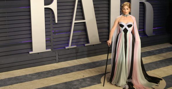 Selma Blair at the 2019 Oscars. She is glamorously dressed in a white, pink & white striped gown and holds a black cane.