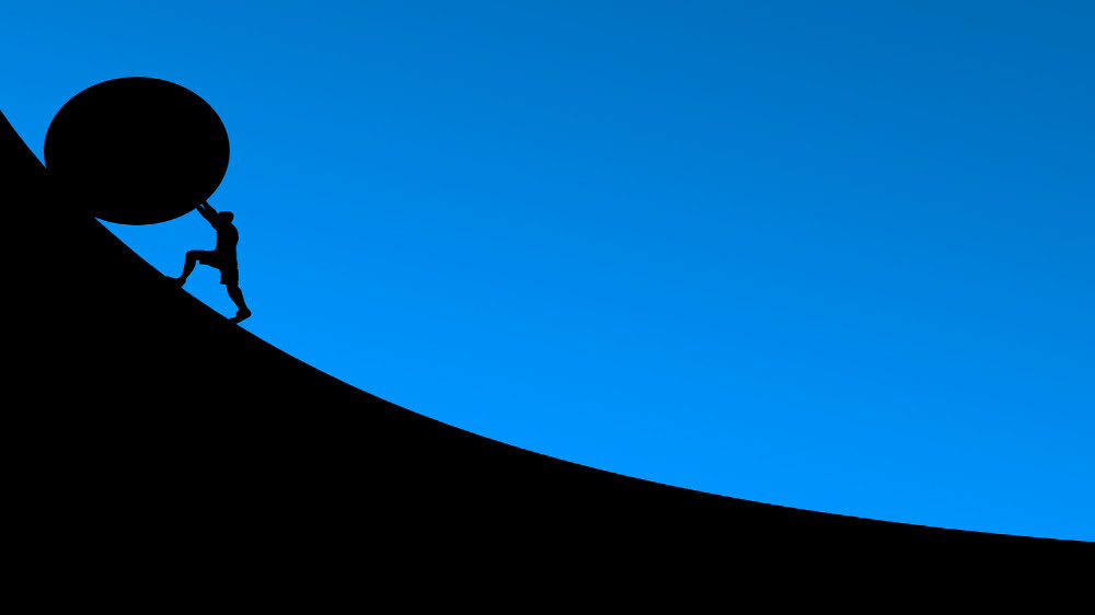 A silhouette of a person pushing a round object that's much bigger than they are up a steep incline.