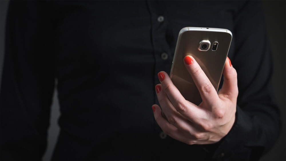 Photo of a person with red-painted fingernails holding a smartphone. On the person's torso/hand is showing.