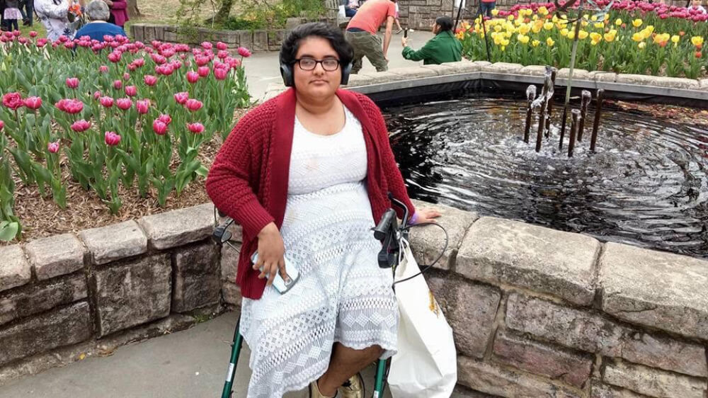 Noor, a south Asian transmasculine person in a white lace dress and red cardigan, sits on a green walker in front of a fountain and a large area of tulips on a sunny day