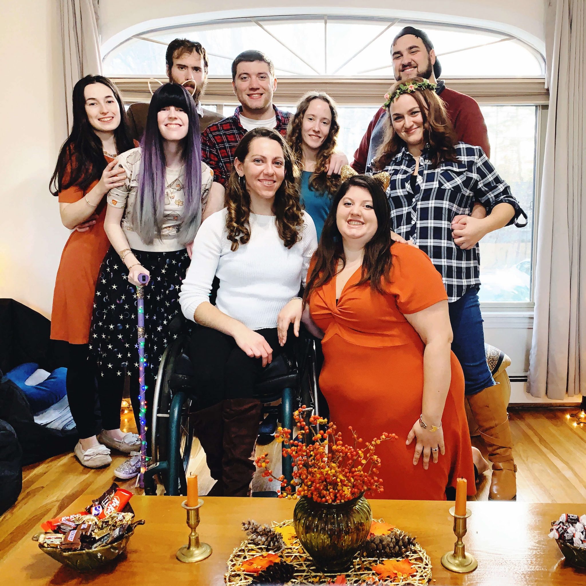The writer is posing with her group of friends for Friendsgiving inside a home with a large window behind everyone. In front, there is a small table with centerpieces and fall-inspired decor, including orange candlesticks. Several people are wearing fall attire, including two orange dresses, a few flannel shirts, a flower crown, a fall sweater with leaves on it, and a purple walking cane decorated with rainbow holiday lights. Everyone is smiling.