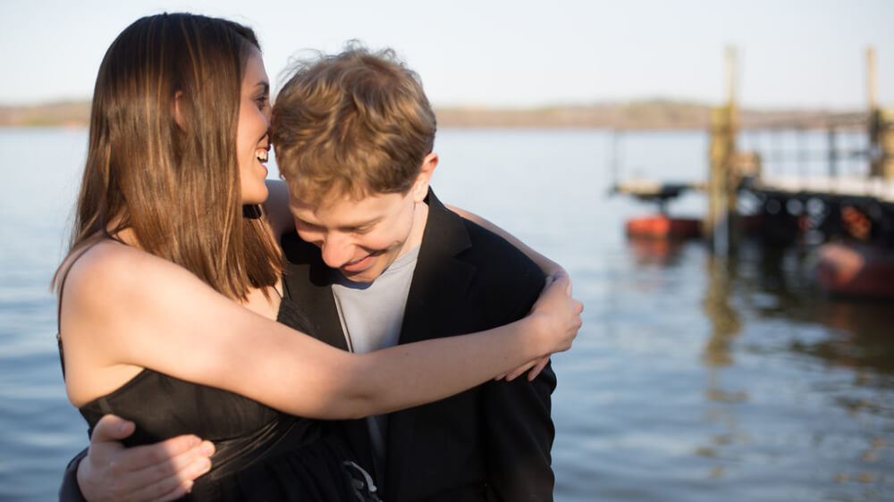 The author and her husband, two smiling white individuals, hug each other in front of a lake. Sarah, who has straight, medium-length brown hair and is wearing a black and white floral dress, speaks into his ear. Her husband, who has wavy, close-cropped dark blonde hair, and is wearing a gray shirt and black suit jacket, bends his head, as if to listen.