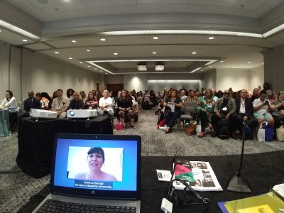 A view of the audience as Rooted in Rights presents at the National Council for Independent Living. On a laptop screen, a disabled parent talks with the audience.