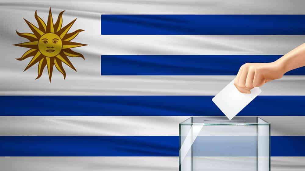 Uruguayan flag with a ballot box in front of it and a hand dropping in the ballot.