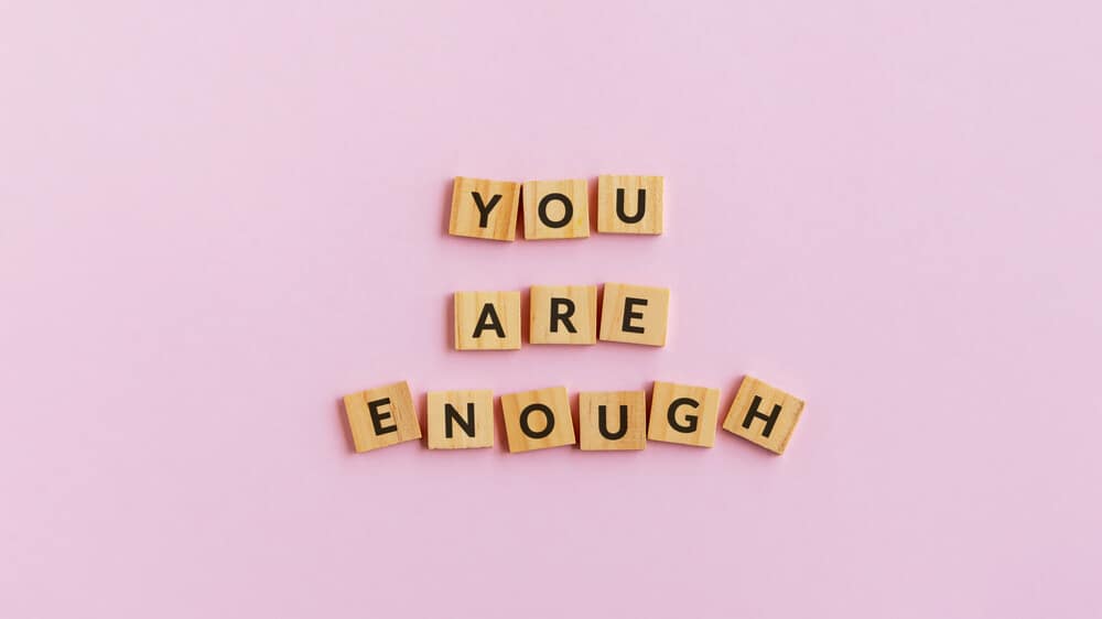 Wooden letter tiles spelling out "you are enough" on a pink background.