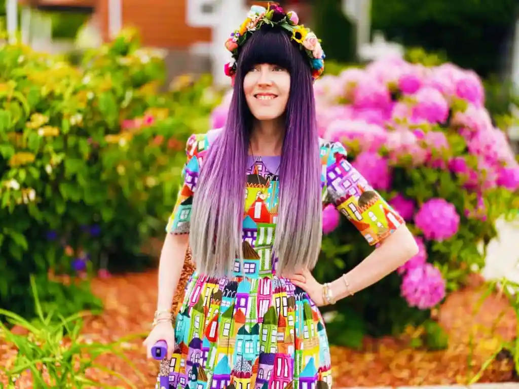 Alaina, a white nonbinary person with long hair that is dark brown fading into lavender and gray, stands outside in front of a colorful garden of flowers. Alaina is wearing a rainbow flower crown on her head and a dress with a colorful houses pattern on it. She has one hand on her hip and another hand on a lavender cane. Alaina has blue eyes that are looking into the distance.