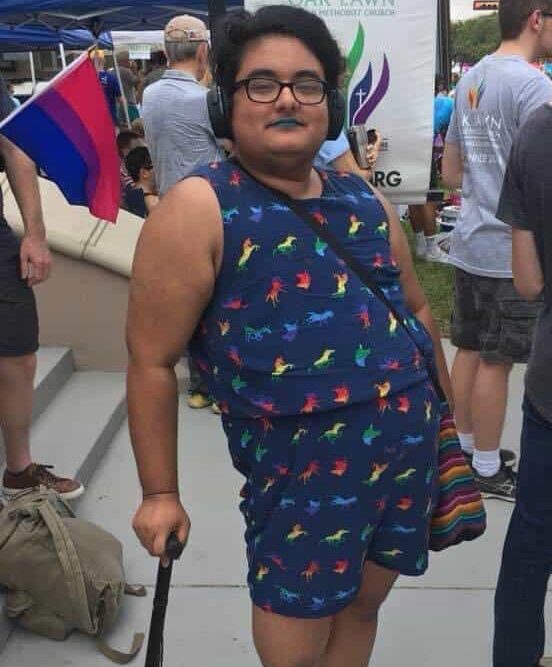 A transmasculine south Asian person leans on a black cane. He wears a rainbow unicorn romper.