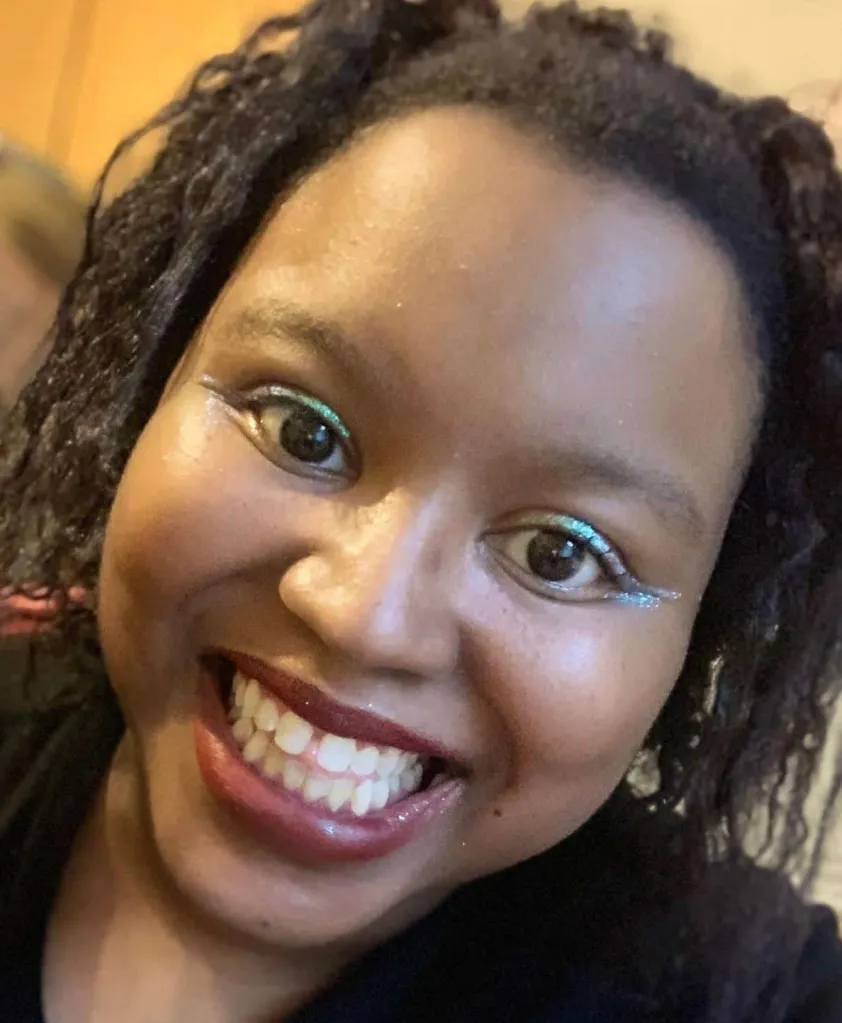 A Black woman, smiling to camera wearing a red lipstick, glitter liner, and a black shirt with white lining