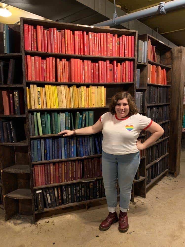 A young woman with short brown hair is standing in front of a tall bookshelf. The books are arranged in rainbow order. She is wearing a white T-shirt with a rainbow heart on the front, jeans, and red boots.