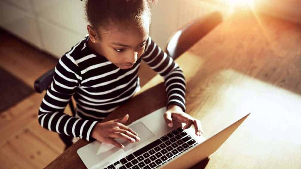 A photo of a young Black child sitting at a desk using a laptop.