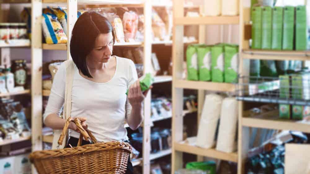 Woman in a grocery store holding a basket in one hand and a product in the other hand, reading its label.