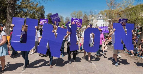 A group of people marching down a street, holding big purple letters that spell "union."