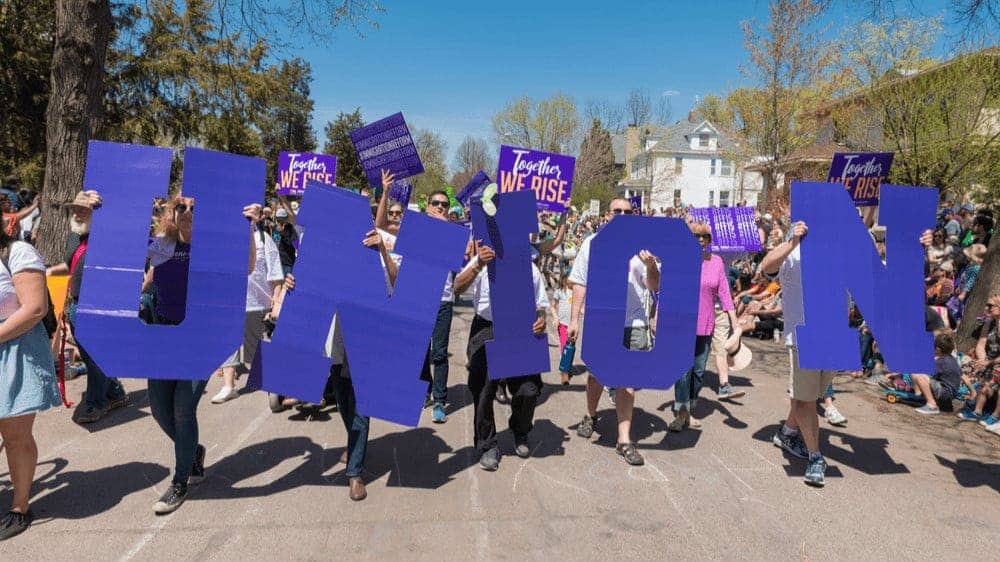 A group of people marching down a street, holding big purple letters that spell "union."